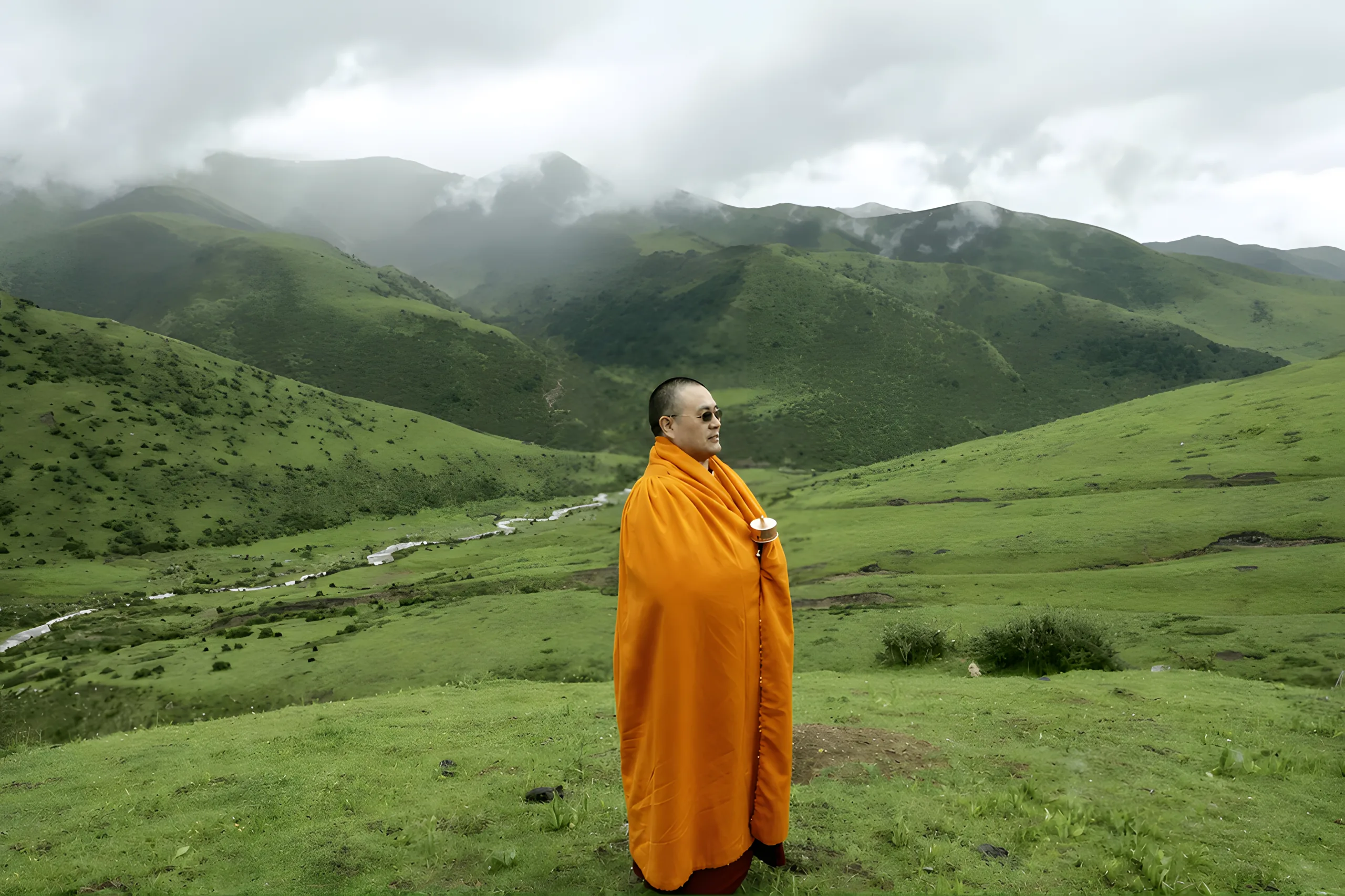 A Buddhist teacher in an orange robe standing in a green mountain valley