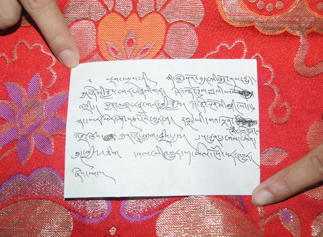 A small handwritten Tibetan prophecy document in black ink rests on red patterned brocade.