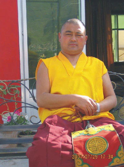 Hor shul Khenpo Gelek Palsang Rinpoche sits outdoors wearing a yellow shirt and red monastic skirt.