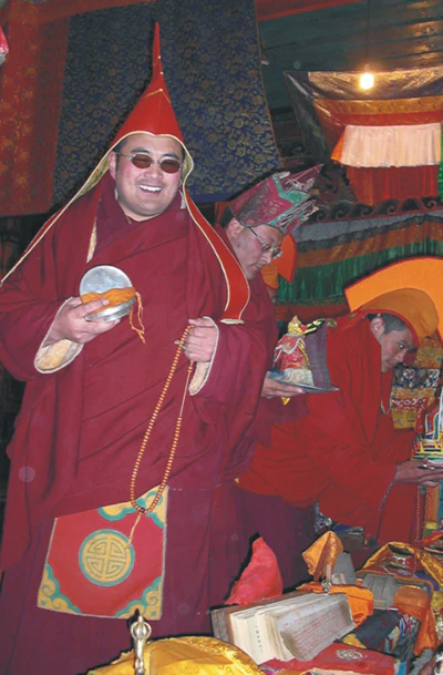 Hor shul Khenpo Gelek Palsang Rinpoche stands in the shrine hall of Hor shul Monastery wearing a red ceremonial hat and holding ritual implements, with monastics behind him.