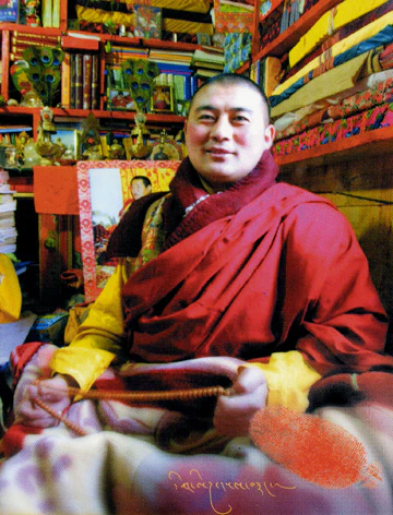 Hor shul Khenpo Gelek Palsang Rinpoche sits holding prayer beads in red and yellow monastic robes, with scriptures and painted woodwork behind him.