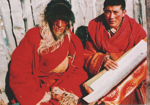 Venerable Qiaq and Hor shul Khenpo Gelek Palsang Rinpoche sit outside a wooden building and examine a long manuscript together.