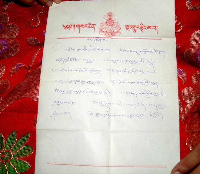 A handwritten Tibetan prophecy document in blue ink with a red printed letterhead rests on red patterned brocade.