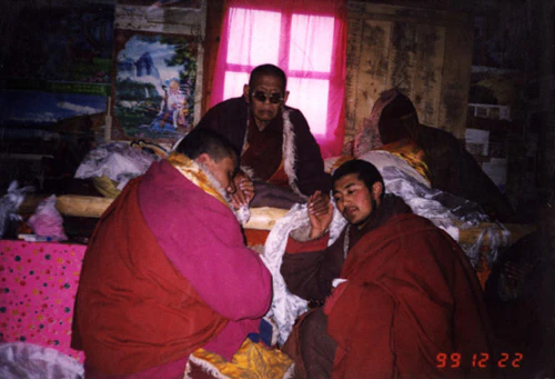 Lama Achuk Rinpoche, Hor shul Khenpo Gelek Palsang Rinpoche, and others speak together beside a bed indoors, wearing monastic robes.
