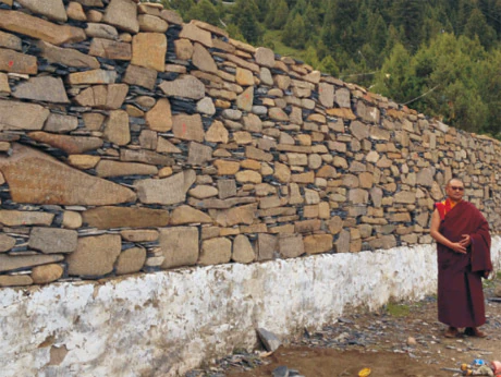 Hor shul Khenpo Gelek Palsang Rinpoche stands in red monastic robes beside a long stone wall at Ala Sacred Mountain.
