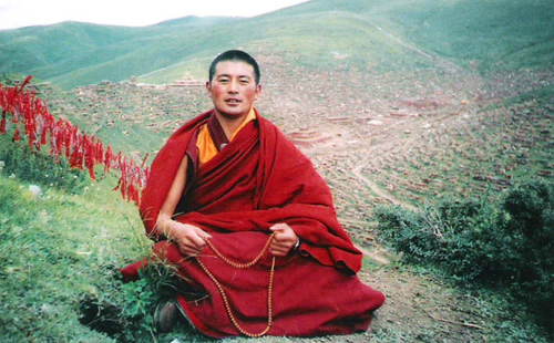 Hor shul Khenpo Gelek Palsang Rinpoche sits on a grassy hillside in red monastic robes, with a dense monastic settlement in the background.