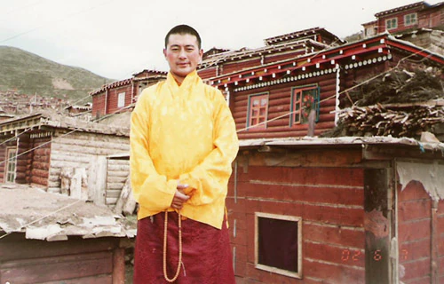 Hor shul Khenpo Gelek Palsang Rinpoche stands in a yellow shirt before the wooden residence where he stayed while teaching at Larung Gar Five Sciences Buddhist Institute.