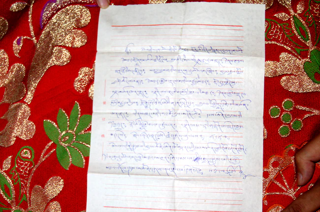 A page of handwritten Tibetan prophecy text in blue ink rests on red patterned brocade.
