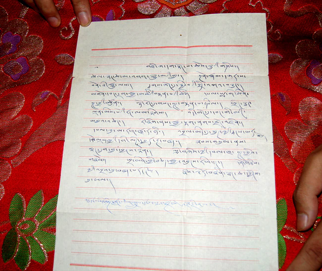 A page of handwritten Tibetan prophecy text in blue ink rests on red patterned brocade, with fingers visible at the right.