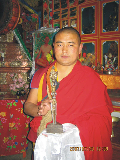 格花堪布身著僧衣，坐在寺院佛堂內，手持金剛橛。