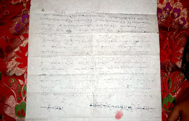 An unfolded gray-white handwritten Tibetan prophecy document rests on red patterned brocade.